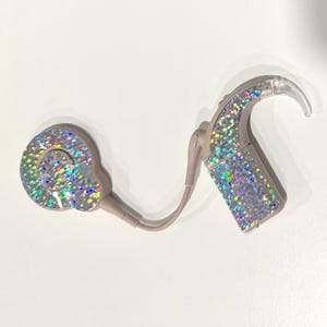 May include: A beige hearing aid with a sparkly, iridescent finish. The device has a curved design with a clear, hook-like component and a circular piece. The hearing aid is connected by a thin, beige wire.