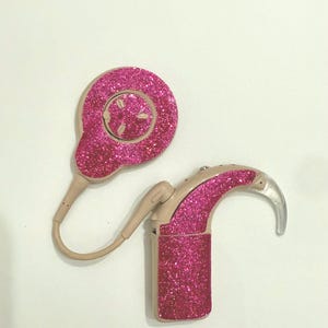 Pode incluir: Dois aparelhos auditivos rosa com glitter. Um tem formato circular com um pequeno botão no centro. O outro tem formato curvo com uma peça de plástico transparente na extremidade. Ambos são bege e rosa.