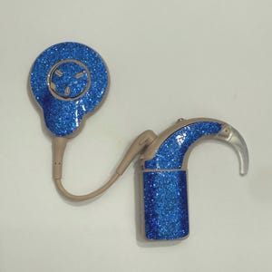 Puede incluir: Un audífono con acabado de purpurina azul. El dispositivo tiene un diseño curvo con una punta plateada y un componente redondo cubierto de purpurina. Un cable beige conecta las dos partes. El componente redondo tiene una cara sonriente.