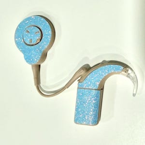 Pode incluir: Um aparelho auditivo azul claro com um acabamento brilhante e cintilante. O aparelho auditivo tem um design curvo com uma ponta transparente e um fone de ouvido circular. O dispositivo é conectado por um fio fino de cor castanho claro.
