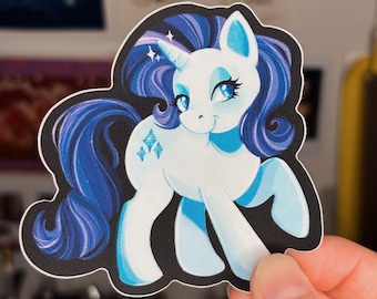 May include: A sticker featuring a white cartoon pony with blue hair and accents. The pony has a unicorn horn and diamond-shaped cutie marks. The sticker is outlined in black and held by a hand.
