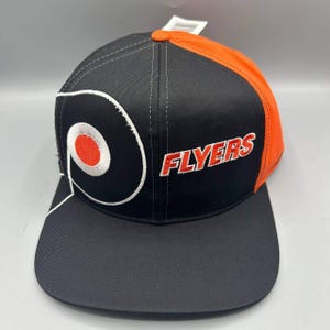 May include: A black and orange baseball cap featuring the Philadelphia Flyers logo embroidered in white and orange on the front panel. The word FLYERS is stitched in orange with a white outline on the side. The cap has a flat brim and a structured crown.