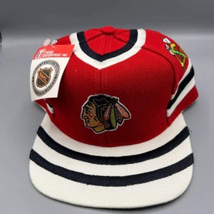 Può includere: Cappellino snapback con licenza NHL dei Chicago Blackhawks, rosso e bianco. Il cappellino presenta il logo della squadra sul davanti, con strisce nere e bianche sulla visiera. Un'etichetta è attaccata.