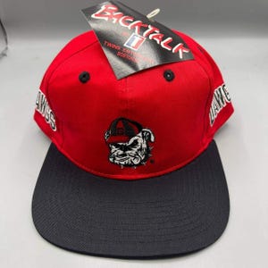 Op de afbeelding: Een rode en marineblauwe baseballcap met het woord "DAWGS" aan de zijkant en een bulldoglogo op de voorkant. De cap heeft een platte klep en een label met de tekst "BACKTALK".