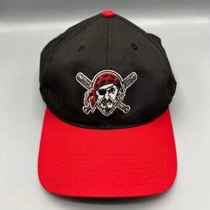 Può includere: Cappellino da baseball nero con visiera rossa e bottone rosso. Il cappellino presenta un logo pirata con un teschio con una bandana rossa e un orecchino d'oro, con mazze da baseball incrociate dietro il teschio.