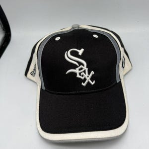 Könnte beinhalten: Eine schwarze Baseballkappe mit weißer und grauer Umrandung. Auf der Vorderseite der Kappe befindet sich das gestickte weiße "SOX"-Logo. Die Kappe hat einen gebogenen Schirm und eine graue Oberseite.