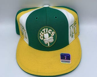 Può includere: Un cappellino da baseball verde, bianco e giallo con la scritta "CELTICS" e un logo a trifoglio. Il cappellino ha "NY" sul lato e un adesivo "FITTED 7". La visiera è gialla.