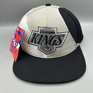 Puede incluir: Una gorra de béisbol con un diseño en gris y negro que presenta el logotipo de los Los Angeles Kings bordado en la parte frontal. El logo incluye un icono de corona y el texto Los Angeles KINGS. Una etiqueta roja y azul de la marca Pro Player está sujeta al lateral.