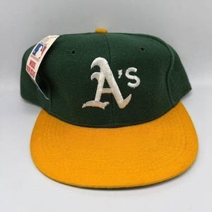 May include: A dark green baseball cap with a yellow brim and the white embroidered A's logo on the front. A white tag attached to the side features the Major League Baseball logo and the text Wool Replicas in red and black lettering.