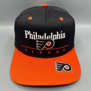Op de afbeelding: Een zwarte honkbalpet met een oranje klep en een oranje knoop aan de bovenkant. De voorkant is voorzien van wit geborduurde tekst Philadelphia, een Flyers-logo en het woord FLYERS in het rood. Een tweede Flyers-logo is op de klep geborduurd.
