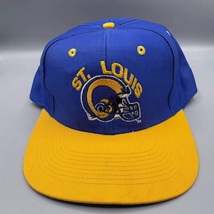 May include: A blue and yellow St. Louis Rams baseball cap. The cap has the team's logo and the words "ST. LOUIS" embroidered in gold. The peak is yellow, and the crown is blue.