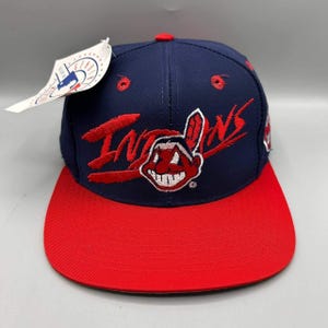 Può includere: Un cappellino da baseball blu navy con visiera rossa e un logo Indians ricamato in rosso raffigurante un personaggio dei cartoni animati. Il cappellino presenta un'etichetta bianca attaccata sul lato con la scritta Genuine Merchandise.