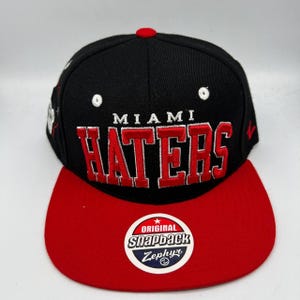 Op de afbeelding: Zwarte en rode snapback-pet met "MIAMI HATERS" in het rood geborduurd. De pet heeft een rode klep, een rode knoop bovenop en een "Snapback Zephyr" patch. Een stijlvol accessoire.