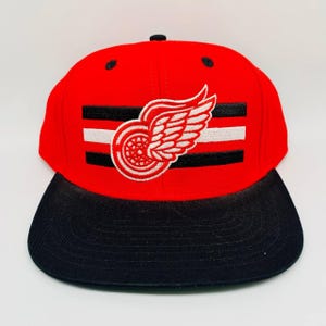 Op de afbeelding: Een rode en zwarte baseballcap met een platte klep. De pet heeft een wit en zwart gestreept ontwerp met een logo van een gevleugeld wiel. De pet heeft een gestructureerde pasvorm en een klassieke, sportieve stijl.
