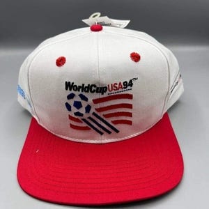 Könnte beinhalten: Weiß-rote Baseballkappe mit dem Text "WorldCup USA94" und einer Fußballgrafik. Die Kappe hat einen roten Schirm und rote Stickereidetails. Ein Etikett ist angebracht.