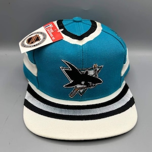 Pode incluir: Um boné de beisebol nas cores azul-petróleo, branco e preto com o logotipo bordado dos San Jose Sharks na frente. O boné tem uma aba reta com listras pretas e prateadas e uma etiqueta NHL Twins Enterprise presa no painel superior.
