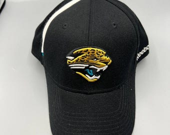 May include: Black baseball cap with a white stripe on the side and a team logo on the front. The logo features a jaguar in gold, teal, and white. The word "Reebok" is embroidered on the side.