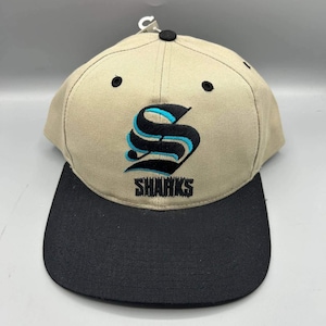 Puede incluir: Una gorra de béisbol de color beige con visera negra y un botón negro en la parte superior. El frente presenta un logotipo bordado de una letra S estilizada en negro y turquesa, con la palabra SHARKS bordada en letras negras debajo.