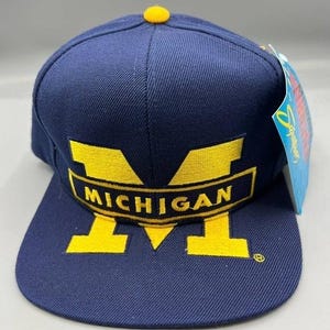 Puede incluir: Gorra de béisbol azul marino con el logotipo "MICHIGAN" bordado en amarillo. La gorra tiene una visera plana, un botón amarillo en la parte superior y una etiqueta adjunta. El logotipo es en negrita.