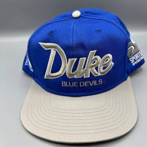 Może przedstawiać: Niebiesko-beżowa czapka z daszkiem z napisem "Duke" wyszytym na biało i szaro. Pod spodem znajduje się napis "BLUE DEVILS". Czapka ma płaski daszek i guzik na górze.
