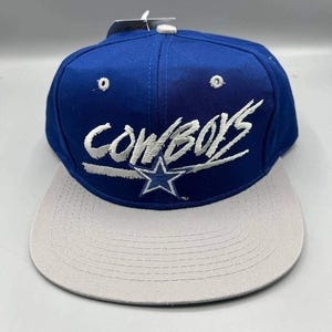 Pode incluir: Boné de beisebol azul e cinza com a palavra "Cowboys" em escrita branca na frente. O boné apresenta um logotipo de estrela e uma aba plana. É um estilo clássico.