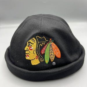 May include: Black skull cap featuring an embroidered logo of a Native American head with yellow, red, and black details, and colorful feathers. The cap is made of a soft, dark fabric.