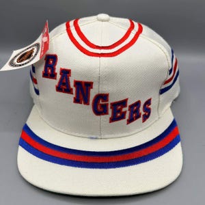 Op de afbeelding: Een witte honkbalpet met het woord RANGERS in blauw borduursel met rode omlijning op de voorzijde. De pet heeft rode en blauwe strepen op de kroon en de klep, en een wit label met het NHL-logo bevestigd aan de zijkant van de pet.
