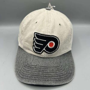 Può includere: Un cappellino da baseball con corona beige chiaro e visiera grigio scuro. La parte anteriore presenta un logo ricamato dei Philadelphia Flyers in nero, bianco e arancione. Il cappellino ha una struttura a sei pannelli con occhielli.