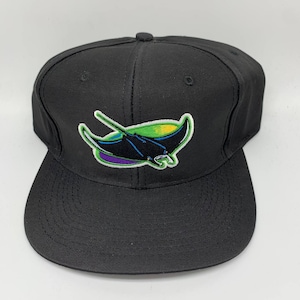 Peut inclure: Casquette de baseball noire avec une visière plate. La casquette présente un écusson brodé d'une raie manta stylisée en noir, vert, jaune et violet. La casquette est fabriquée dans un tissu durable et a une coupe structurée.