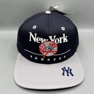 Puede incluir: Una gorra de béisbol azul marino con visera plana gris claro. El frente presenta texto bordado en blanco que dice New York y Yankees, con un gráfico central de béisbol que contiene la palabra Yankees. Un logo NY azul marino está en la visera.