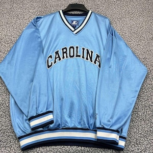 Op de afbeelding: Lichtblauwe Starter-trui met een V-hals. Het woord "CAROLINA" staat in zwarte letters in een boog over de borst. De manchetten en zoom hebben strepen in wit, marineblauw en lichtblauw.