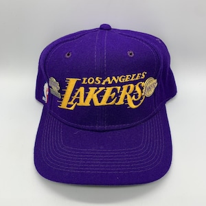 May include: Purple baseball cap with the words "Los Angeles Lakers" embroidered in yellow. The cap has a curved brim and a structured crown. The Lakers logo is prominently displayed on the front.