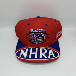 May include: A red and blue baseball cap with the text "Speed Limit 325 mph" and "NHRA" embroidered on the front. The cap has a flat brim and a structured crown, with a small logo on the side.