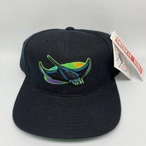 Op de afbeelding: Zwarte baseballcap met een platte klep, met een geborduurd manta-rogontwerp in blauw, groen en paars. De cap heeft een label met de tekst "AMERICAN NEEDLE".