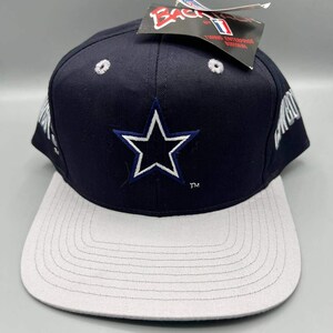 Op de afbeelding: Een marineblauwe en grijze baseballcap met een witte ster en blauwe omtrek aan de voorkant. De cap heeft een platte klep en het woord "Cowboys" is aan de zijkant zichtbaar. Er is een label aan de bovenkant bevestigd.
