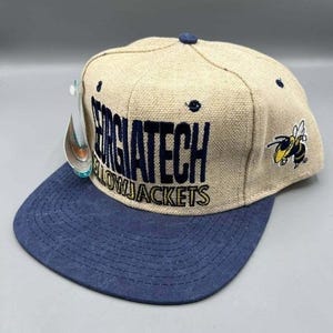 Puede incluir: Gorra de béisbol beige y azul marino con las palabras "GEORGIA TECH YELLOWJACKETS" en letras de bloque azul marino. Un logotipo de abeja amarilla y negra está en el lateral. La gorra tiene una visera plana y un botón en la parte superior.