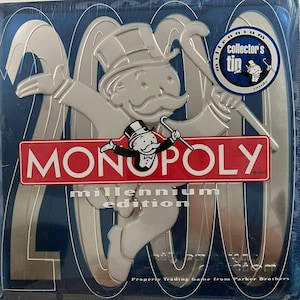 Puede incluir: Juego de mesa Monopoly Millennium Edition en una lata azul y plateada. El juego presenta el logotipo de Monopoly en rojo y blanco, con el icónico hombre de Monopoly. La lata tiene un diseño plateado de 2000 y una pegatina de consejo para coleccionistas.