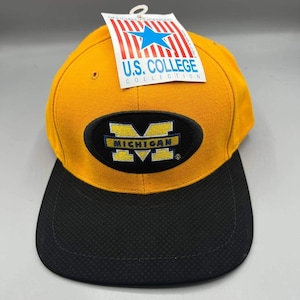 Puede incluir: Gorra de béisbol amarilla y negra con la palabra "MICHIGAN". La gorra tiene una visera negra y una etiqueta que dice "U.S. COLLEGE COLLECTION". Es una gorra de estilo snapback.