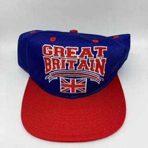 May include: A blue and red baseball cap with the words "GREAT BRITAIN" in white block letters. The cap features the Union Jack flag and a red brim. The cap is a classic style.