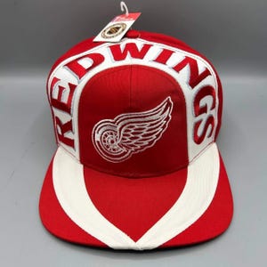 Pode incluir: Um boné de beisebol vermelho e branco com o logótipo dos Detroit Red Wings bordado a branco no painel frontal. A palavra RED WINGS está cosida em letras brancas grandes na coroa. Uma etiqueta branca da NHL está presa ao topo.