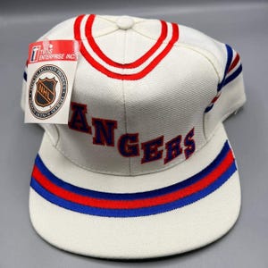 Puede incluir: Gorra de béisbol blanca con la palabra "RANGERS" en letras azules. La gorra presenta rayas rojas y azules en el borde y una raya roja alrededor de la corona. Una etiqueta de Twins Enterprise Inc. está adjunta.