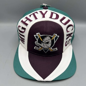 May include: A teal, white, and dark purple baseball cap featuring the embroidered text MIGHTY DUCKS in dark purple lettering. The centre of the cap displays an embroidered logo of a duck goalie mask crossed with two hockey sticks in white, yellow, and grey.