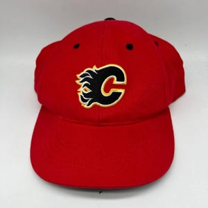 Może przedstawiać: Czerwona czapka bejsbolowa z logo Calgary Flames. Logo to czarna litera 'C' z płomieniem i żółtymi akcentami. Czapka ma zakrzywiony daszek i czarny guzik na górze.