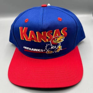 May include: A blue and red Kansas Jayhawks baseball cap. The cap features the word "Kansas" in yellow, with a Jayhawk mascot logo. The brim is red, and the top of the cap is blue.