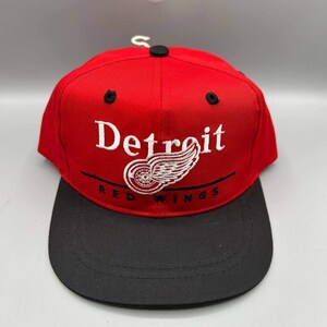 Puede incluir: Una gorra de béisbol roja con visera negra y un botón negro en la parte superior. El frente presenta el texto bordado en blanco Detroit sobre un logotipo de rueda alada blanca, con el texto negro RED WINGS bordado debajo del logo.