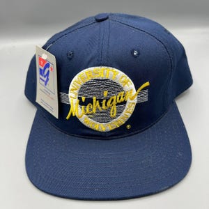 May include: Navy blue baseball cap with a flat brim. The cap features a circular emblem with the words "UNIVERSITY OF Michigan WOLVERINES" in yellow and white. A white tag is attached to the side.