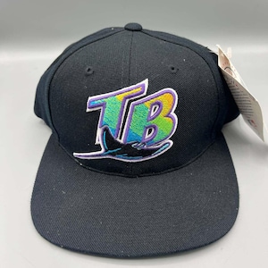 Puede incluir: Gorra de béisbol negra con el logotipo de los Tampa Bay Rays. El logotipo incluye las letras "TB" en verde azulado, morado y amarillo, con una manta raya estilizada. La gorra tiene una visera plana y una etiqueta adjunta.