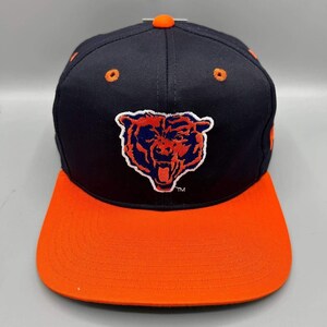 May include: A navy blue baseball cap featuring an embroidered orange bear head logo on the front panel. The cap has a flat orange brim, an orange top button, and orange eyelets. The bear logo is outlined in white with a small TM symbol below it.