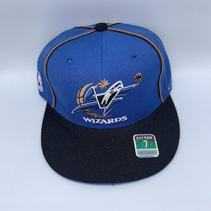 Puede incluir: Gorra de béisbol de los Wizards azul y negra con el logotipo del equipo bordado en la parte delantera. La gorra tiene una visera negra y una pegatina de talla 7. El logotipo presenta un baloncesto y una estrella.