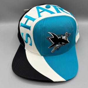 Puede incluir: Una gorra de béisbol con una combinación de colores turquesa, blanco y negro. El panel frontal muestra un logotipo de tiburón bordado con un palo de hockey. La palabra SHARKS está bordada en turquesa en los paneles laterales blancos.